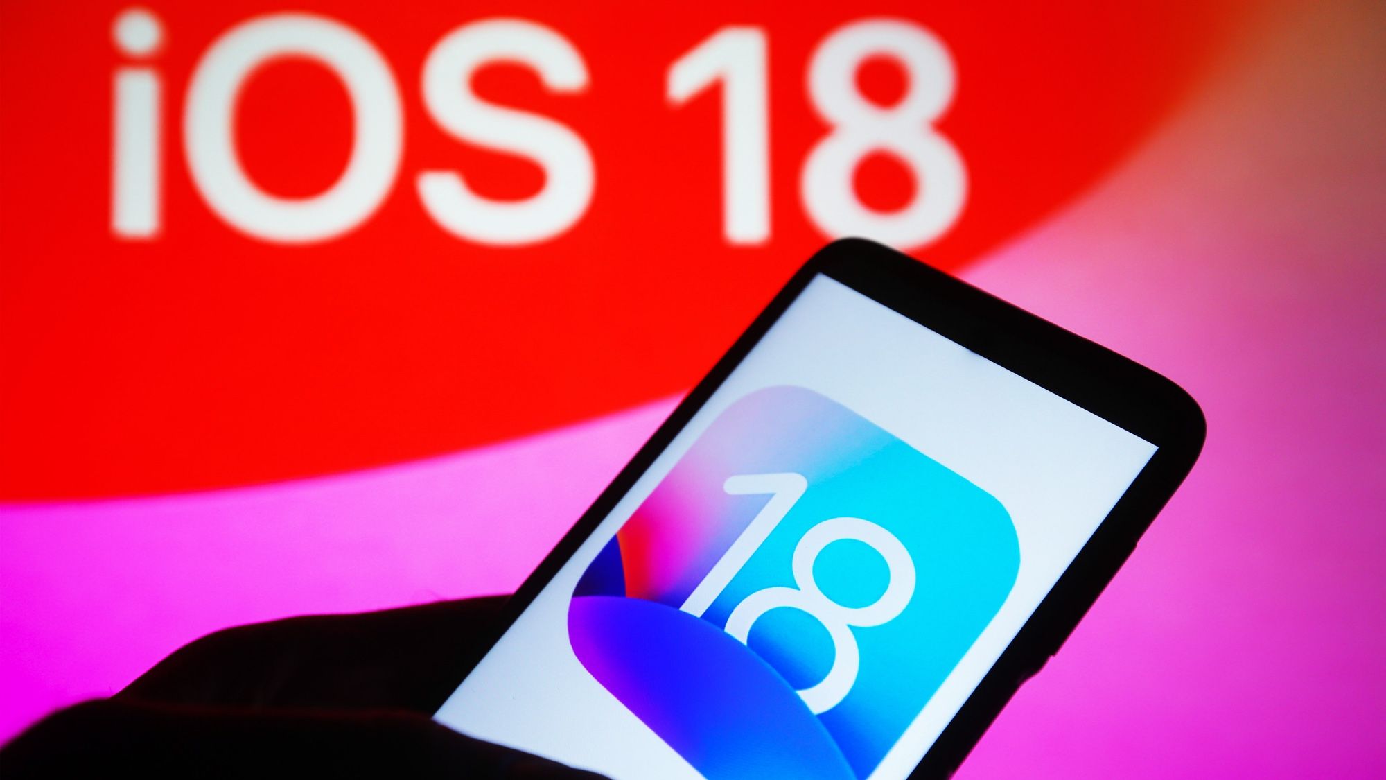 Mão esquerda a segurar um telemóvel cujo ecrã tem um quadrado estilizado com o número 18 no centro. Atrás, lê-se "iOS 18".