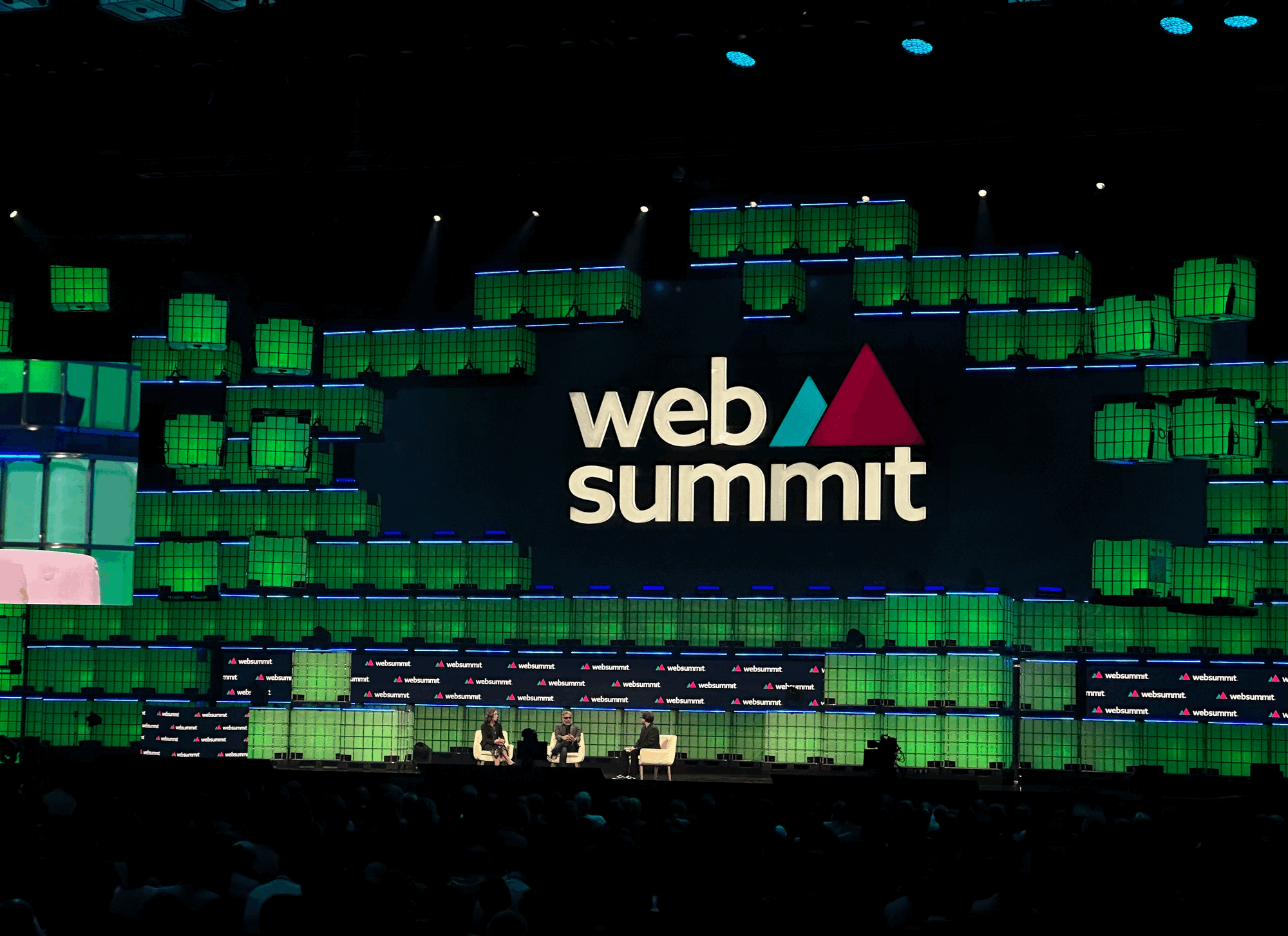Palco principal da Web Summit 2023 visto ao longe com o logo em destaque. No palco, Julie Pace, Yasir Khan e David Meyer.