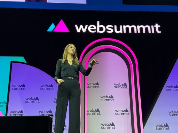 Daniela Braga no palco 13 da Web Summit 2023 com um comando na mão direita. Acima, o logo da conferência.