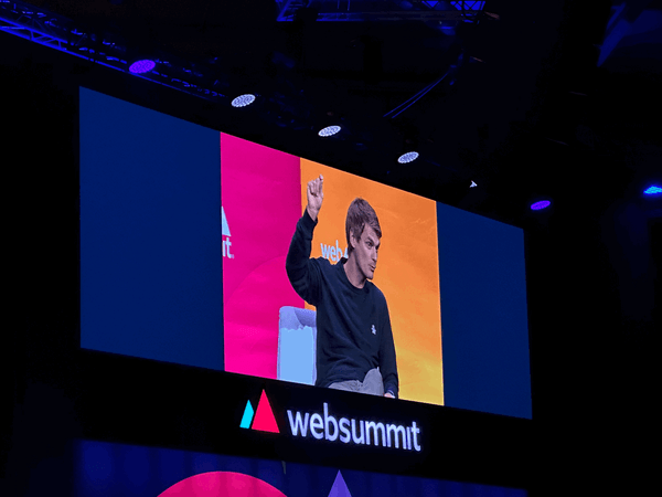 Sebastian Weidt no ecrã do palco 11 da Web Summit 2023, com a mão no ar. Abaixo, o logo da conferência.